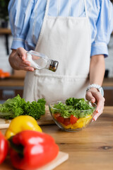 partial view of young adult woman in apron seasoning fresh vegetables salad with salt mill in modern kitchen