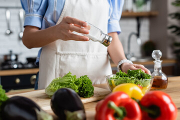 partial view of young adult woman in apron seasoning fresh vegetables salad with salt mill in kitchen