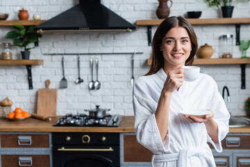 smiling young adult woman in bathrobe drinking coffee and looking at camera in modern loft