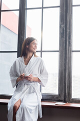 pretty young adult woman in bathrobe sitting with cup of coffee near window at home