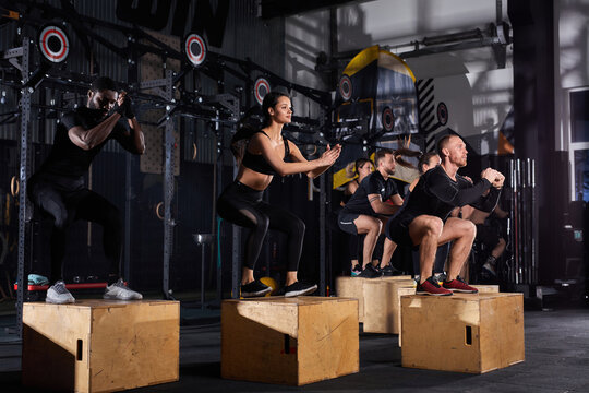 Side View On People Jumping On Wooden Box At Gym. Young People Performing Intense Workout Routine, In Sportswear. Sport, Cross Fit concept