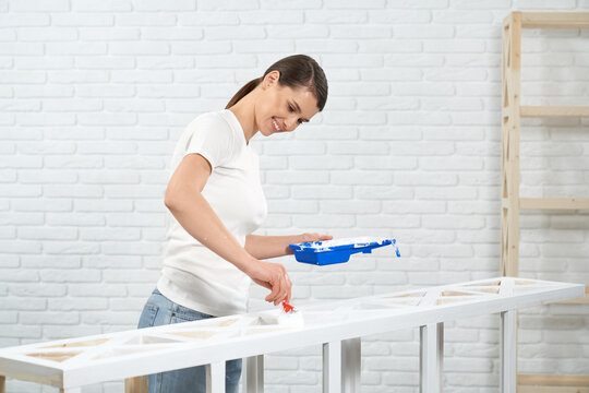 Smiling brunette in casual outfit painting wooden storage stand in white color with brush. Happy woman refreshing old shelves at home. Working process.