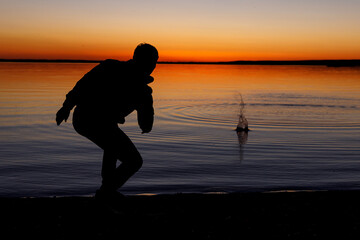 Silhouette of a man throws stones into the lake at sunset
