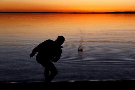 Silhouette of a man throws stones into the lake at sunset