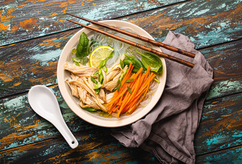 Asian soup with rice noodles, chicken and vegetables in ceramic bowl served with spoon and...