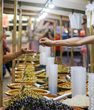 A Man’s Arm Reaches Out To Give Change Back To A Woman Buying Olives At The Market In Mallorca. 