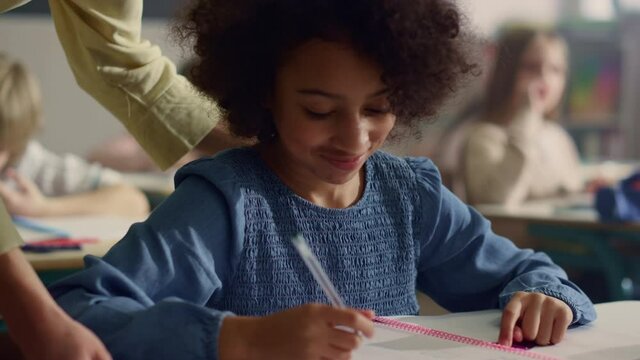 Girl talking with teacher in classroom. Smiling schoolgirl writing in notebook