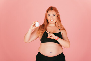 Fototapeta na wymiar Happy plus size woman wearing sporty outfit pointing at jar of pills