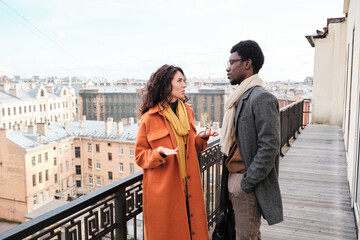 Obraz premium Young multiethnic couple in coats standing on balcony and talking