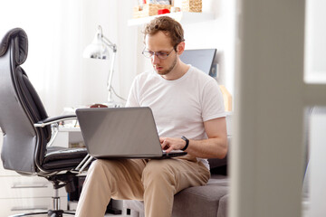 Young man with beard, working at home in living room sitting on coach with his laptop in front of him.
