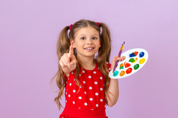 A little girl with curly hair holds a paint palette and points her finger forward.