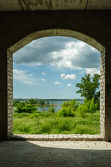 A view of the river, green grass, and sunny skies through an archway in an abandoned brick house.