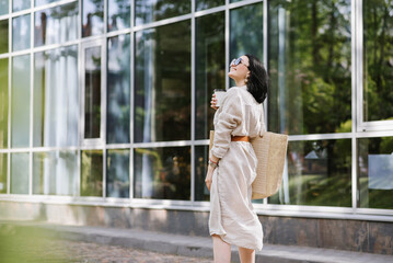 Brunette young woman with sunglasses and bag holding coffee walking in the city. Lifestyle portrait of woman