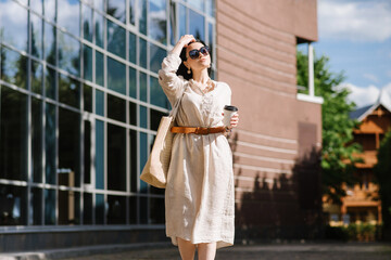 Brunette young woman with sunglasses and bag holding coffee walking in the city. Lifestyle portrait of woman