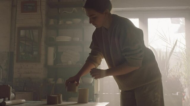 Craftswoman is standing at workplace in workshop in counter light, cutting clay with string tool into smaller pieces, working with pleasure and inspiration, Slow motion.