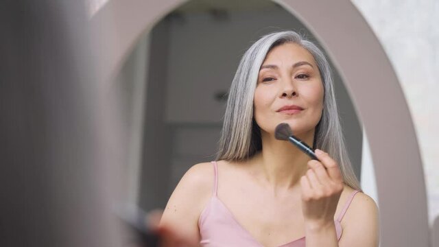 Senior older middle aged Asian woman with grey hair holding make up brush on radiant face with perfect skin looking at mirror smiling to her reflexion. Ads of makeup foundation for natural glow skin.
