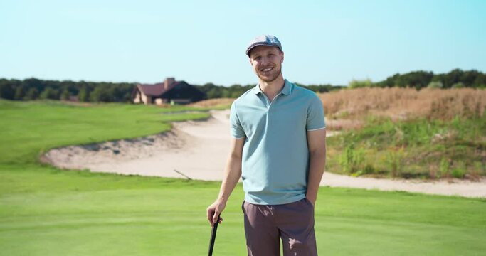 Portrait of positive male caucasian golf player smiling, standing with golf club at golf course