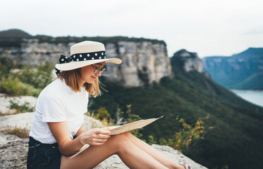 tourist girl in hat holding in hands looking on map resting during summer trip in mountains landscape, hipster woman in glasses plans route relax in nature outdoors, empty space for text