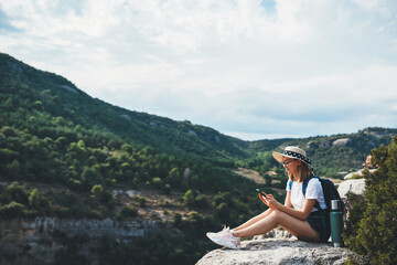 cute girl traveler in hat resting during vacation in nature and plans route online map in smartphone, tourist girl uses mobile phone while enjoy vacation on  background of panorama mountain landscape