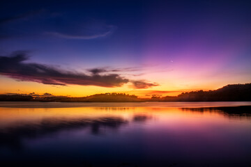 Fototapeta na wymiar Uma paisagem focada no pôr-do-sol com montanhas e um lago espelhado e cristalino com cores vibrantes azul, amarelo, magenta e laranja com algumas nuvens contrastantes com a paisagem.