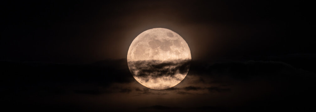 horizontal panoramic image of the full moon slightly obscured by clouds during the night