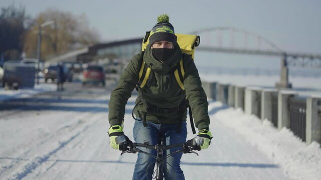 Portrait of cyclist with backpack and protective mask rides bicycle on snow covered street with food delivery. Man works as courier and delivers order to customers during virus outbreak. Slow motion.
