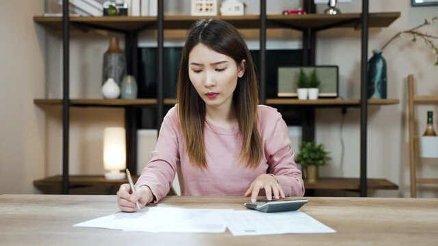 Stressed Asian woman looking at the paper bills calculating credit card invoice or unpaid taxes and bank account overpay at home