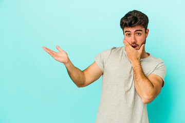 Young caucasian man isolated on blue background holds copy space on a palm, keep hand over cheek. Amazed and delighted.