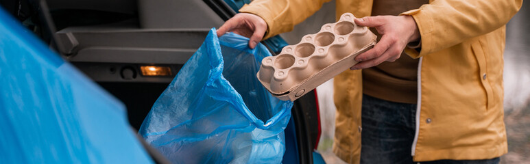 cropped view of man holding carton container near blue trash bag in car, banner