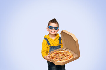 Little cute baby boy in a yellow sweater and denim overalls and blue sunglasses smiles and holds an open box with pizza isolated on background.