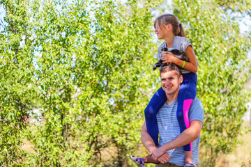 Naklejka na ściany i meble Defocused brother riding sister on back. Portrait of happy girl on man shoulders, piggyback. Girl holding cat, kitten. Family playing outdoor. Love animal. Green background. Copy space. Out of focus