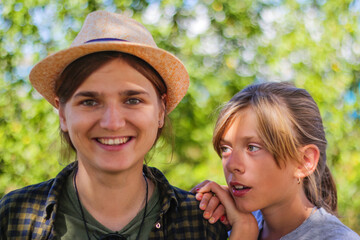 Defocused close-up smiling young brunette caucasian woman in hat with preteen girl. Summer blurred green nature bokeh background. Portrait of handsome young cute woman and teen girl. Out of focus