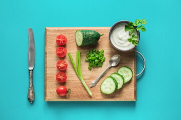 Fresh vegetables for salad, cucumbers, cherry tomatoes, green onions and white yoghurt on a wooden cutting board on a blue background. Top view, copy space.