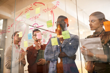 Pensive start-up team brainstorming in front of mind map on glass wall while working on new...