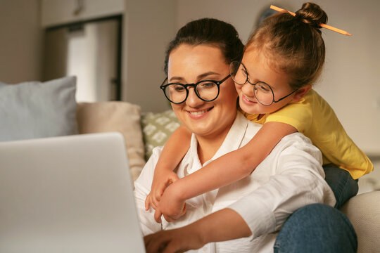 Young Happy Woman Mother In Glasses Surfing Online On Laptop With Little Girl Daughter, Excited Child Hugging Smiling Mom While Spending Time Together In Light Cozy Living Room At Home