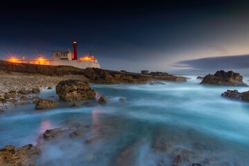 Seascape night long exposure, rocky beach on foreground and lighthouse and streaky clouds on background, Colour Photo, Lisboa, Portugal