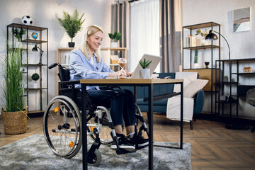 Fototapeta na wymiar Handicapped woman with blond hair using wireless laptop for remote work from home. Pretty female sitting in wheelchair and typing on keyboard.