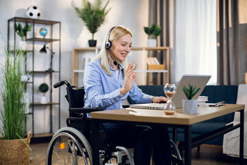 Pretty young woman in wheelchair using headset and laptop for video conference at home. Handicapped...