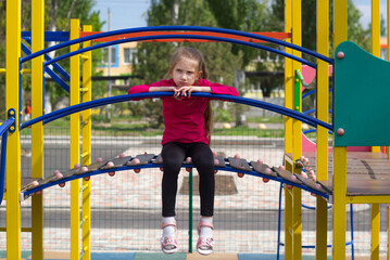 Obraz premium A child girl with blond hair in a pink T-shirt is sad on the playground, sits on a slide, her legs dangling. 