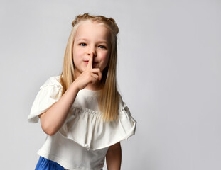 Children's secret. Close up portrait of a little blonde girl showing a gesture of silence on a white background. Cute baby makes a quiet gesture. Child holds a finger to his mouth shows a sign of Shh