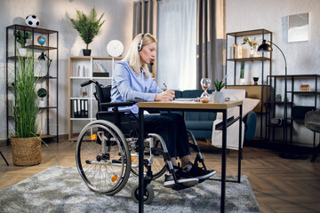 Side view of woman with disabilities taking notes during video conference at home. Female...