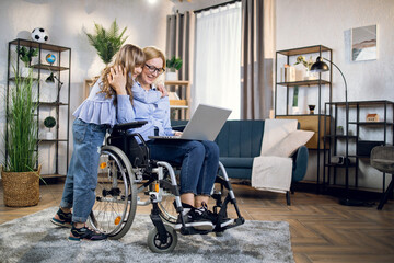 Charming woman in wheelchair working on laptop while her cute daughter standing near and hugging her. Disabled mother with child at home. - obrazy, fototapety, plakaty