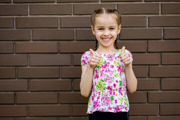 The little girl smiles and shows two thumbs up. A beautiful child stands against the background of a brick wall with a place for text. Emotional girl is dressed in bright clothes and is very happy