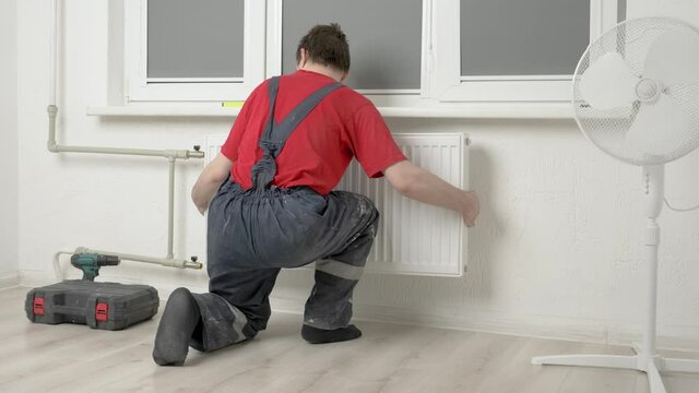 The plumber installs a white metal radiator to the central heating pipes in the room, there are white walls and windows, the concept of construction and heating maintenance