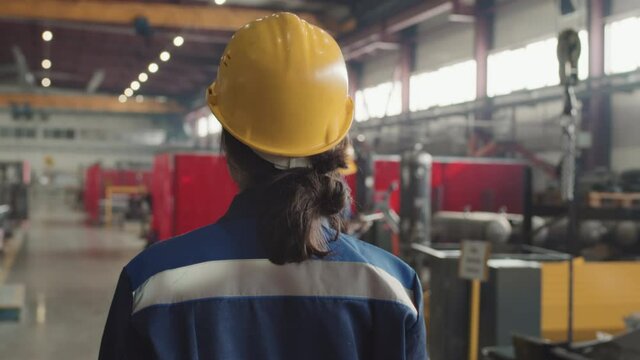 Rear-view tracking slowmo of female supervisor in hard hat and uniform walking through plant facility controlling work of employees