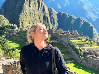 Naklejka premium Blond woman smiling and relaxing in ancient fortress Machu Picchu Peru on sunny summer day. Happy woman in ancient Inca lost city Machu Picchu. Happiness, adventure, dream, smile.