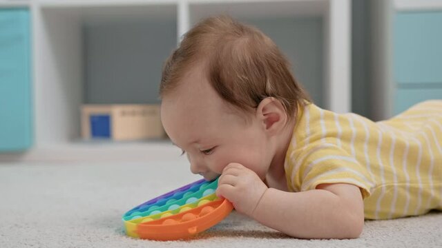 Child plays with anti-stress toy popping bubbles with his finger and chewing on silicone toy with his mouth. Newborn three-month-old baby plays with colorful rainbow pop it toy while lying on carpet.