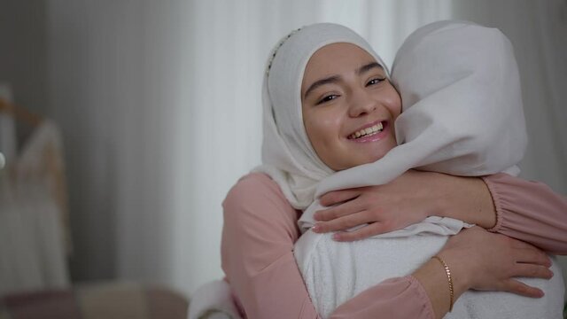 Happy Middle Eastern bridesmaid in hijab hugging bride smiling looking at camera. Positive young charming Muslim woman supporting friend getting married on wedding morning indoors