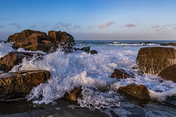 Closeup of foamy wave rushing ashore amid rockly coast of Laguna Beach, California. Early morning sky, clouds in the distance. 
