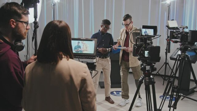Full shot of backstage of filmmaking process in studio. Male operator with leg prosthesis and African American presenter discussing storyboard while video assistant and gaffer adjusting equipment